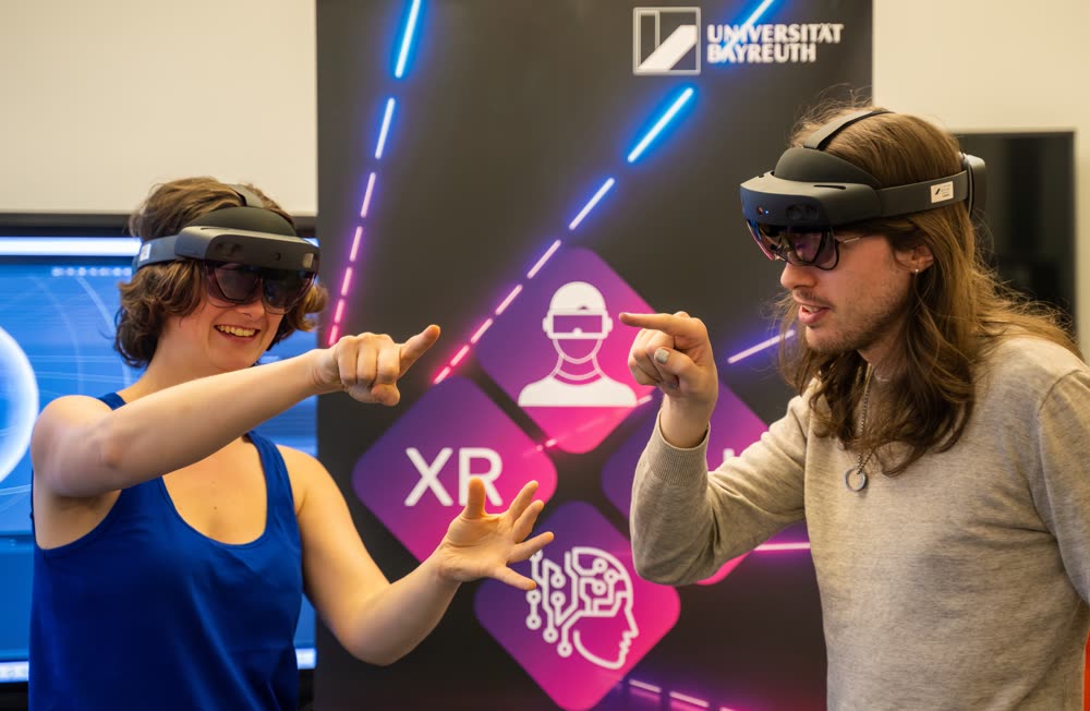Zwei Personen probieren HoloLens-Brillen aus und zeigen lachend aufeinander, im Hintergrund ein XR-Banner der Universität Bayreuth